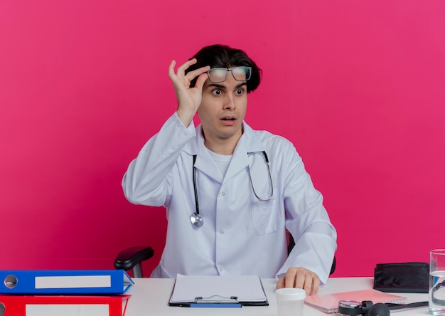 Impressionato giovane medico maschio che indossa veste medica e stetoscopio con gli occhiali seduto alla scrivania con strumenti medici guardando i vetri afferranti laterali isolati sul muro rosa