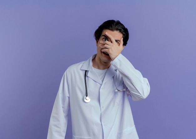 Impressionato giovane medico maschio che indossa veste medica e stetoscopio mettendo la mano sul viso attraverso le dita isolate sulla parete viola con lo spazio della copia