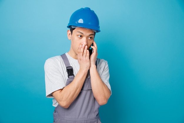 Impressionato giovane operaio edile che indossa il casco di sicurezza e sussurra uniforme sul telefono guardando a lato