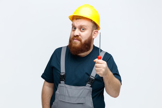 Impressionato giovane operaio edile maschio che indossa casco di sicurezza e uniforme guardando la fotocamera tenendo il cacciavite isolato su sfondo bianco