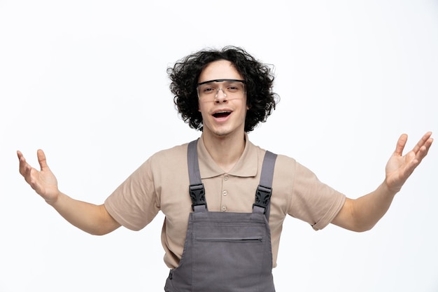 Impressionato giovane operaio edile maschio che indossa uniforme e occhiali di sicurezza guardando la fotocamera che mostra un gesto di benvenuto con le braccia aperte isolate su sfondo bianco