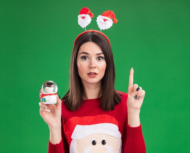 Impressionato giovane ragazza abbastanza caucasica che indossa il maglione di Babbo Natale e fascia che tiene la figurina del pupazzo di neve rivolta verso l'alto isolata sulla parete verde