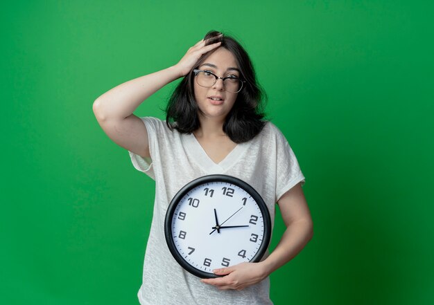 Impressionato giovane ragazza abbastanza caucasica con gli occhiali che tengono l'orologio e mettendo la mano sulla testa isolata su priorità bassa verde con lo spazio della copia