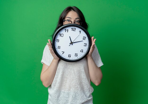Impressionato giovane ragazza abbastanza caucasica con gli occhiali tenendo l'orologio e guardando la telecamera da dietro isolato su sfondo verde con spazio di copia