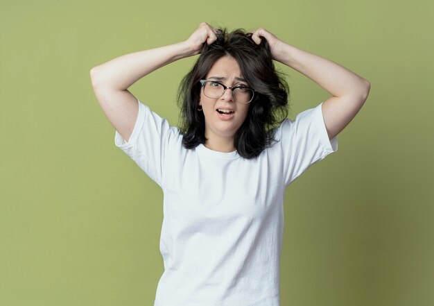 Impressionato giovane ragazza abbastanza caucasica con gli occhiali tirando i capelli isolati su sfondo verde oliva