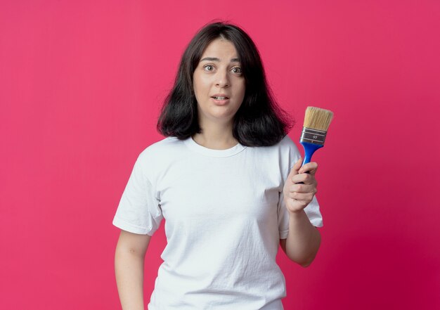 Impressionato giovane ragazza abbastanza caucasica tenendo il pennello isolato su sfondo cremisi con spazio di copia