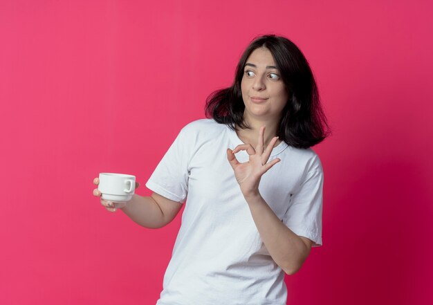 Impressionato giovane ragazza abbastanza caucasica tenendo la tazza guardando a lato e facendo segno ok isolato su sfondo cremisi con lo spazio della copia