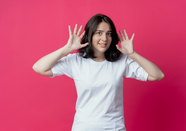 Impressionato giovane ragazza abbastanza caucasica tenendo le mani vicino alla testa isolata su sfondo cremisi