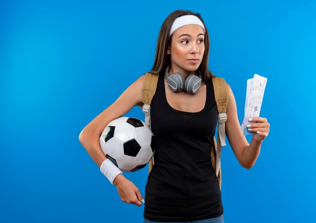 Impressionato giovane ragazza abbastanza sportiva che indossa fascia e cinturino e borsa posteriore con le cuffie sul collo che tiene i biglietti dell'aereo con pallone da calcio isolato sulla parete blu con spazio di copia