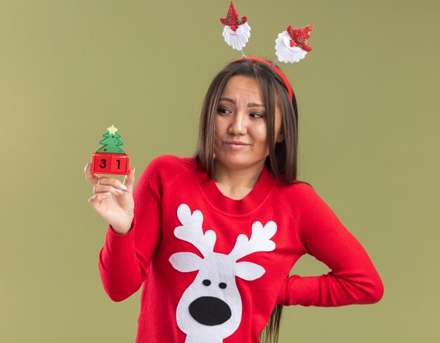 Impressionato giovane ragazza asiatica che indossa il cerchio dei capelli di Natale che tiene e guardando il giocattolo di Natale che mette la mano sull'anca isolata su fondo verde oliva