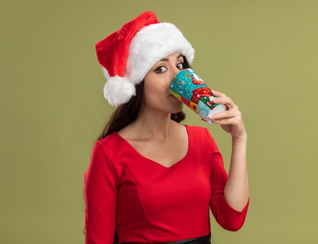 impressionato giovane ragazza carina che indossa il cappello santa tenendo la tazza di caffè di Natale guardando la fotocamera bere caffè isolato su sfondo verde oliva