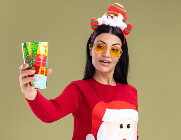 Impressionato giovane ragazza caucasica che indossa la fascia di Babbo Natale e un maglione con gli occhiali che si estende in plastica tazza di Natale guardandolo isolato su sfondo verde oliva
