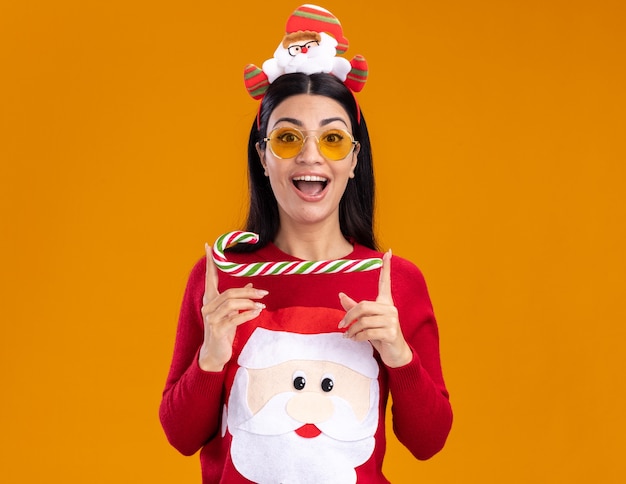 impressionato giovane ragazza caucasica che indossa la fascia di babbo natale e un maglione con gli occhiali che tengono il tradizionale bastoncino di zucchero natalizio isolato orizzontalmente sulla parete arancione con spazio di copia