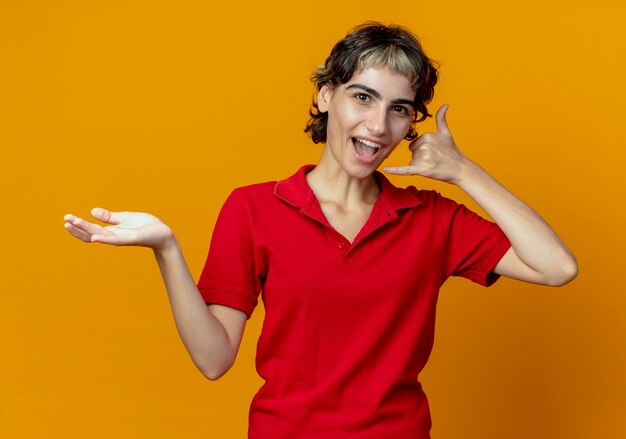 Impressionato giovane ragazza caucasica con taglio di capelli pixie che mostra la mano vuota e facendo gesto di chiamata isolato su sfondo arancione