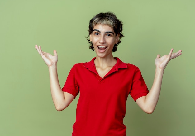 Impressionato giovane ragazza caucasica con taglio di capelli pixie che mostra le mani vuote isolate su sfondo verde oliva con lo spazio della copia