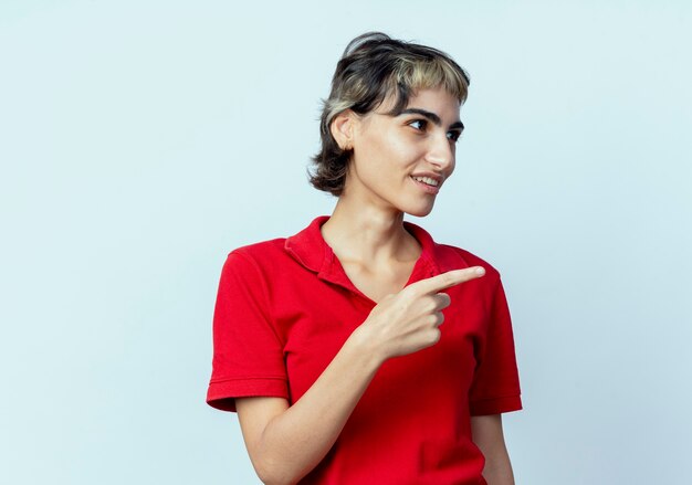 Impressionato giovane ragazza caucasica con taglio di capelli pixie guardando e indicando lato isolato su sfondo bianco con spazio di copia