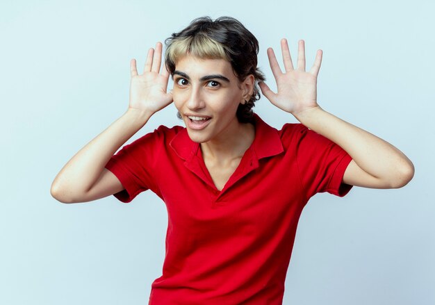 Impressionato giovane ragazza caucasica con taglio di capelli pixie mantenendo le mani vicino alle orecchie isolate su priorità bassa bianca