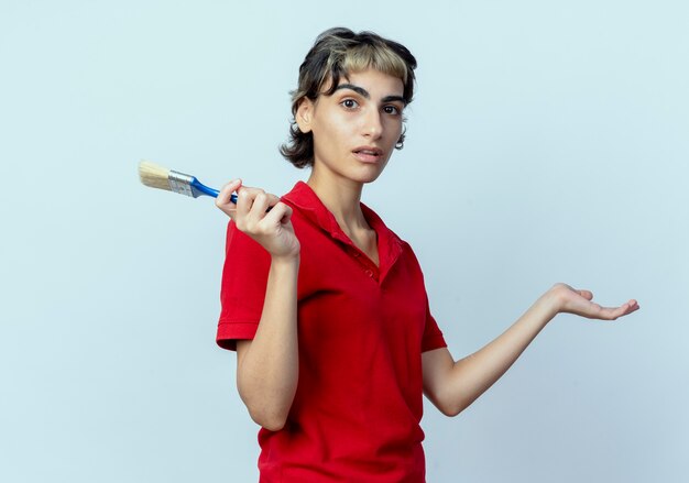 Impressionato giovane ragazza caucasica con taglio di capelli pixie tenendo il pennello che mostra la mano vuota isolata su sfondo bianco