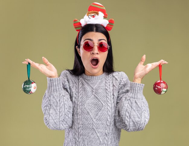 Impressionato giovane ragazza caucasica indossando la fascia di Babbo Natale con gli occhiali che tengono le bagattelle di Natale che guarda l'obbiettivo isolato su sfondo verde oliva