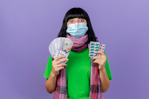 Impressionato giovane ragazza caucasica malata che indossa maschera e sciarpa che tiene soldi e confezioni di capsule guardando la telecamera isolata su sfondo viola con spazio di copia