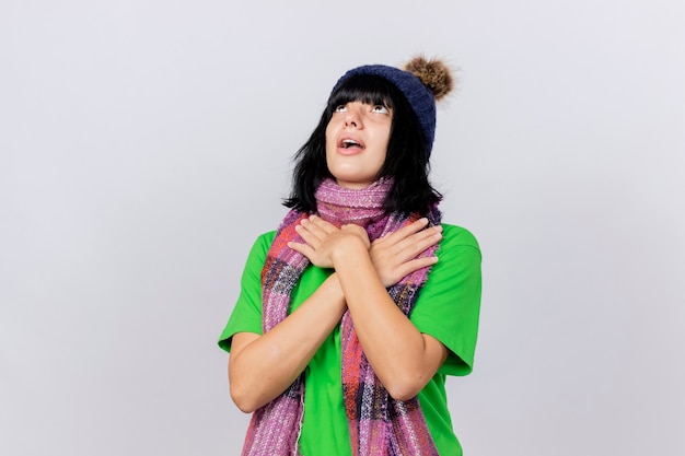 Impressionato giovane ragazza caucasica malata indossando cappello invernale e sciarpa alzando lo sguardo tenendo le mani incrociate sulle spalle isolate su sfondo bianco con spazio di copia
