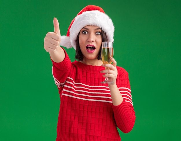 Impressionato giovane ragazza che indossa il cappello di Babbo Natale tenendo un bicchiere di champagne guardando la telecamera che mostra il pollice in alto isolato su sfondo verde