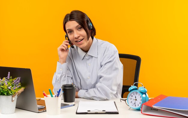 Impressionato giovane ragazza della call center che indossa la cuffia avricolare che si siede allo scrittorio che mette la mano sulla cuffia avricolare isolata sull'arancio