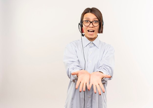 Impressionato giovane ragazza della call center con gli occhiali e auricolare che allunga le mani mostrando le mani vuote isolate su bianco con lo spazio della copia