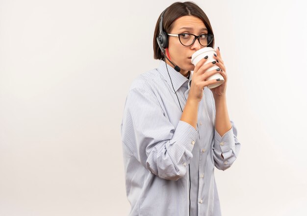 Impressionato giovane ragazza della call center con gli occhiali e auricolare che beve caffè dalla tazza di caffè in plastica guardando il lato isolato su bianco con lo spazio della copia