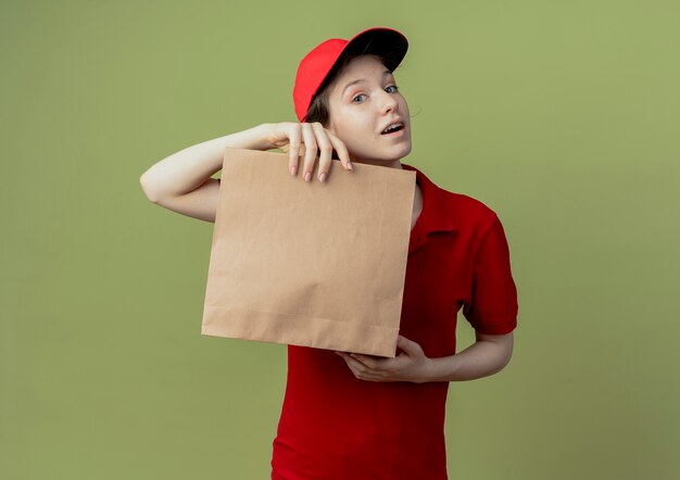 Impressionato giovane ragazza graziosa di consegna in uniforme rossa e cappuccio che tiene il pacchetto di carta che guarda l'obbiettivo isolato su fondo verde oliva con lo spazio della copia