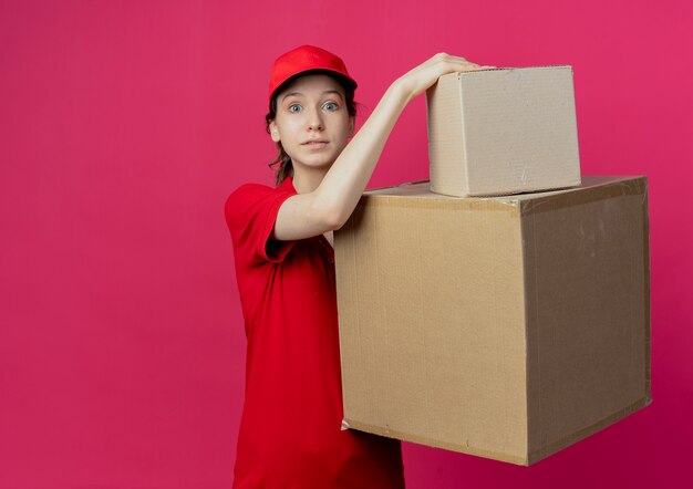 Impressionato giovane ragazza graziosa di consegna in uniforme rossa e cappuccio che tiene le scatole di cartone che guarda l'obbiettivo isolato su sfondo cremisi con lo spazio della copia