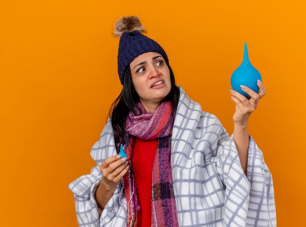 Impressionato giovane ragazza malata caucasica che indossa cappello invernale e sciarpa avvolti in clisteri di contenimento plaid guardando quello grande isolato sulla parete arancione con lo spazio della copia