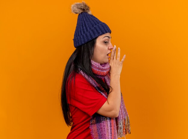 Impressionato giovane ragazza malata caucasica che indossa cappello invernale e sciarpa in piedi in vista di profilo tenendo la mano vicino alla bocca guardando il sussurro laterale isolato sulla parete arancione con lo spazio della copia