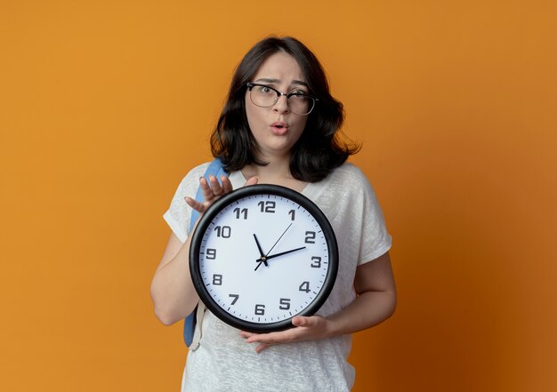 Impressionato giovane ragazza piuttosto caucasica con gli occhiali e borsa posteriore che tiene orologio isolato su sfondo arancione con spazio di copia