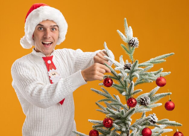 Impressionato giovane ragazzo bello che indossa cappello di Natale e cravatta di Babbo Natale in piedi vicino all'albero di Natale decorandolo con ornamento della palla di Natale che guarda l'obbiettivo isolato su priorità bassa arancione