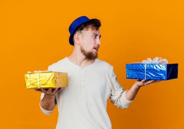 Impressionato giovane ragazzo slavo bello del partito che indossa il cappello del partito che tiene e che esamina i contenitori di regalo isolati su fondo arancio