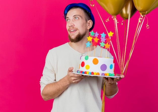 Impressionato giovane ragazzo slavo bello del partito che indossa il cappello del partito che tiene i palloncini e la torta di compleanno con le stelle che esaminano il lato isolato sulla parete rosa con lo spazio della copia