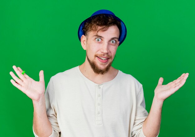 Impressionato giovane ragazzo slavo bello del partito che indossa il cappello del partito guardando la parte anteriore che mostra le mani vuote isolate sulla parete verde