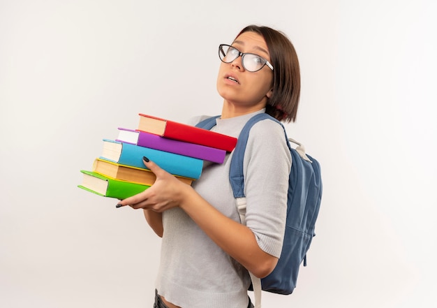 Impressionato giovane studente ragazza con gli occhiali e borsa posteriore in piedi in vista di profilo tenendo libri isolati su bianco