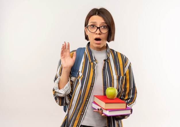 Impressionato giovane studente ragazza con gli occhiali e borsa posteriore in possesso di libri e mela su di loro alzando la mano isolata su bianco