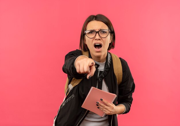 Impressionato giovane studente ragazza con gli occhiali e borsa posteriore tenendo il blocco note che punta alla parte anteriore isolato sul rosa