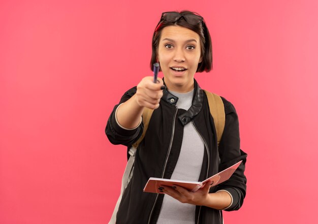Impressionato giovane studente ragazza con gli occhiali sulla testa e sul retro della borsa che tiene il blocco note che indica davanti con la penna isolata sul rosa