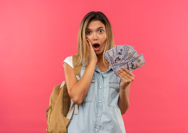 Impressionato giovane studentessa graziosa che indossa la borsa posteriore tenendo i soldi e mettendo la mano sulla guancia isolata sul rosa