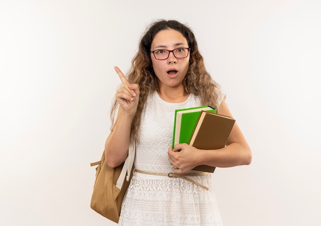 Impressionato giovane studentessa graziosa con gli occhiali e borsa posteriore che tengono i libri che alzano il dito isolato su bianco