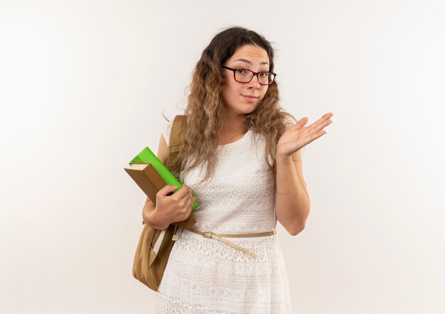 Impressionato giovane studentessa graziosa con gli occhiali e borsa posteriore che tengono i libri che mostrano la mano vuota isolata sopra