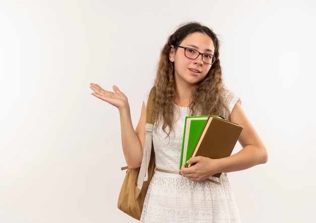 Impressionato giovane studentessa graziosa con gli occhiali e borsa posteriore che tengono i libri che mostrano la mano vuota isolata su bianco