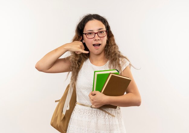 Impressionato giovane studentessa graziosa con gli occhiali e borsa posteriore che tengono i libri facendo gesto di chiamata isolato su bianco