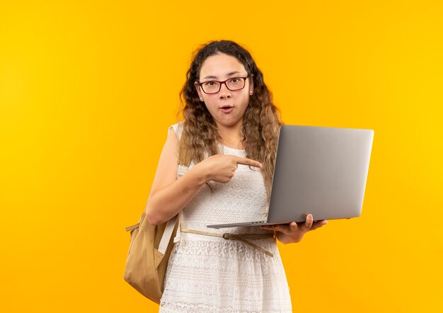 Impressionato giovane studentessa graziosa con gli occhiali e borsa posteriore che tiene e che indica al computer portatile isolato su giallo