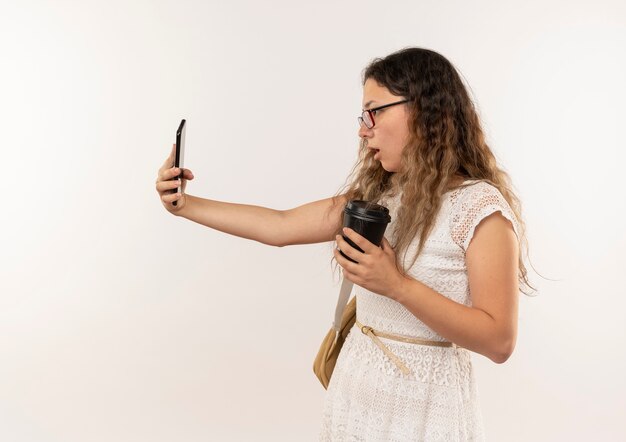 Impressionato giovane studentessa graziosa con gli occhiali e borsa posteriore che tiene e guardando il telefono cellulare con la tazza di caffè in plastica in mano isolato