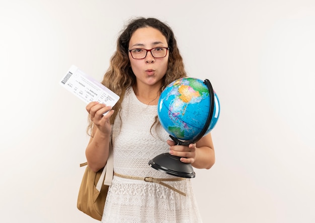 Impressionato giovane studentessa graziosa con gli occhiali e borsa posteriore in possesso di biglietti e globo isolato su bianco